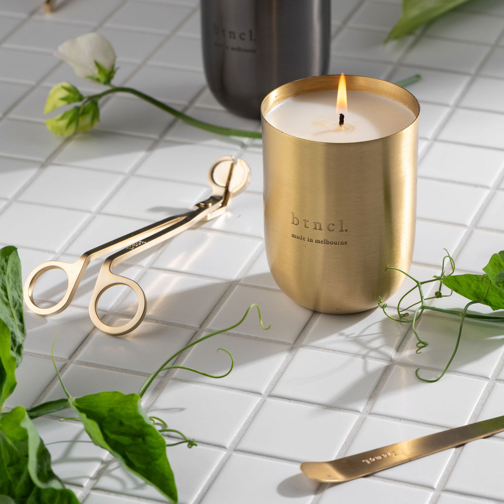 Black and brass scented candle on white tiled surface with botanicals
